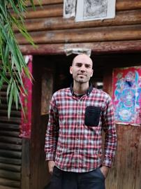 Plan américain. Le sujet de la photographie se trouve face à l'entrée d'une maison en bois, en chemise de flanelle.